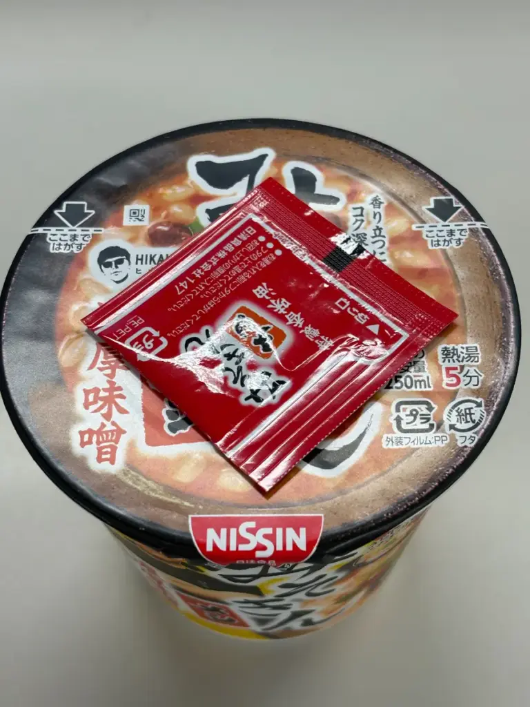 新みそきんメシのパッケージ上部に、赤い特製香味油の小袋が置かれている様子。フタには「熱湯5分」「250ml」と調理方法が記載されている。